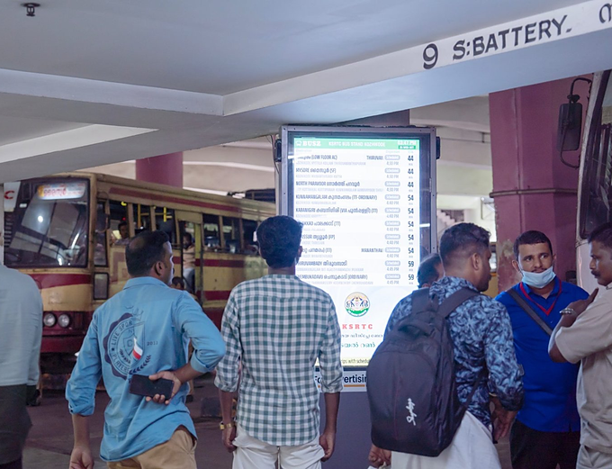 Commuters seated beside a Routerise screen in a bus stand waiting area